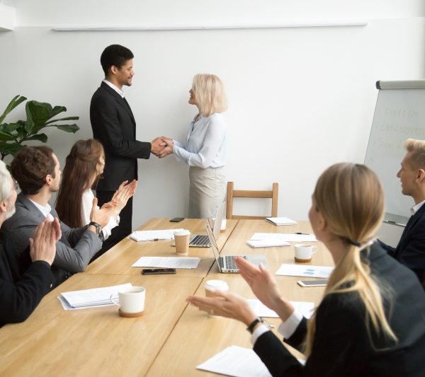 Senior female boss promoting male african employee while team applauding congratulating, aged businesswoman executive and black businessman shaking hands at group meeting, appreciation or recognition