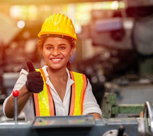 Girl teen worker with safety helmet show thumb up working as labor in industry factory with steel machine.